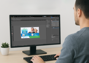 Designer editing a dental clinic postcard with promotional messaging on a widescreen monitor.