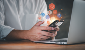 Person holding smartphone in front of laptop with floating social media icons indicating engagement