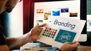 Person reviewing a brand board with color palette, font styles, and logo drafts on paper in front of a desktop.