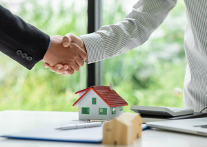 Close-up of handshake at a real estate closing table with model home and signed documents.